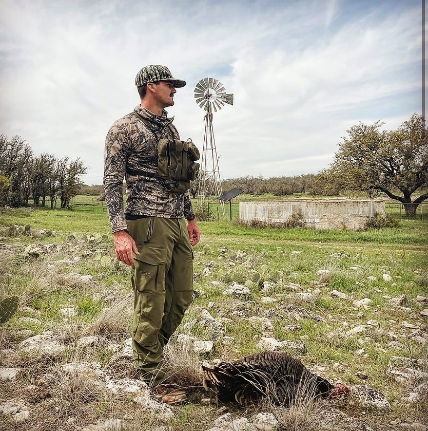 A man in camo and green pants standing above a freshly hunted turkey against a cloudy blue sky