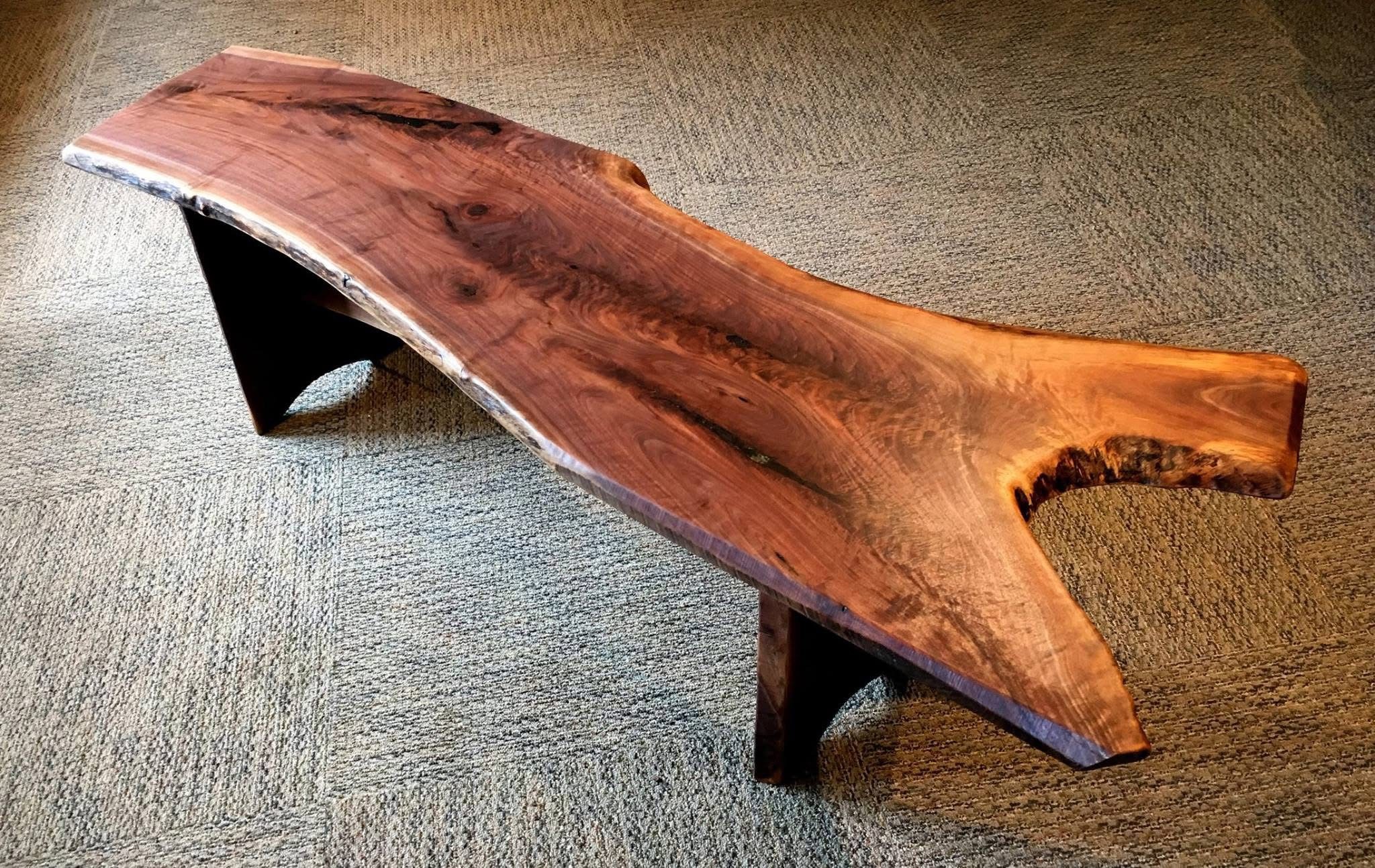 A black walnut live edge bench with wooden legs, one end of the bench is straight, while the other end has a "crotch" section of the tree